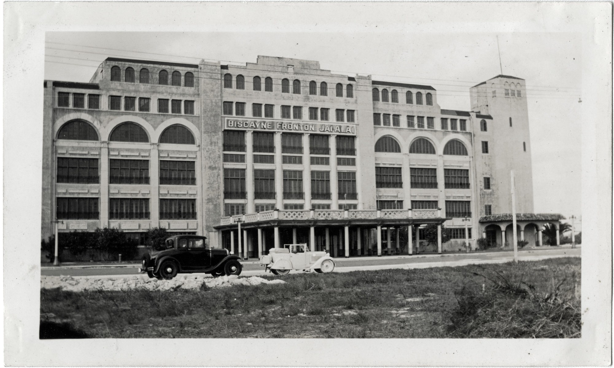 The Biscayne Fronton Jai Alai in Miami, a five-story Mediterranean-revival building with the words "Biscayne Fronton Jai Alai" on the facade and two 1920s automobiles parked in front.