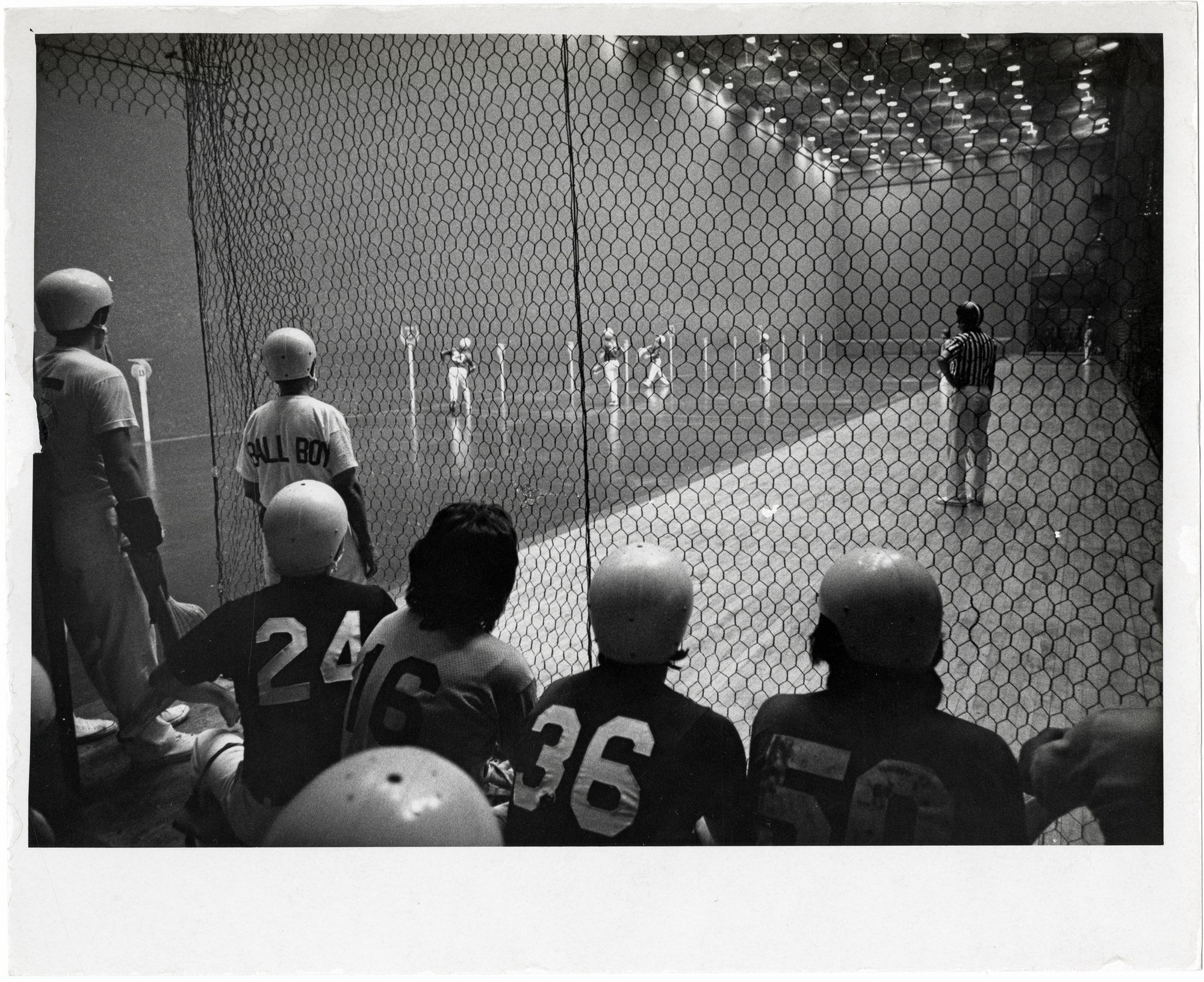 Helmeted pelotaris waiting on the bench behind the safety mesh at a Florida Jai Alai fronton during the Golden Age; a "BALL BOY" assistant, a striped-shirt referee, and players lined up between numbered posts on the granite court.