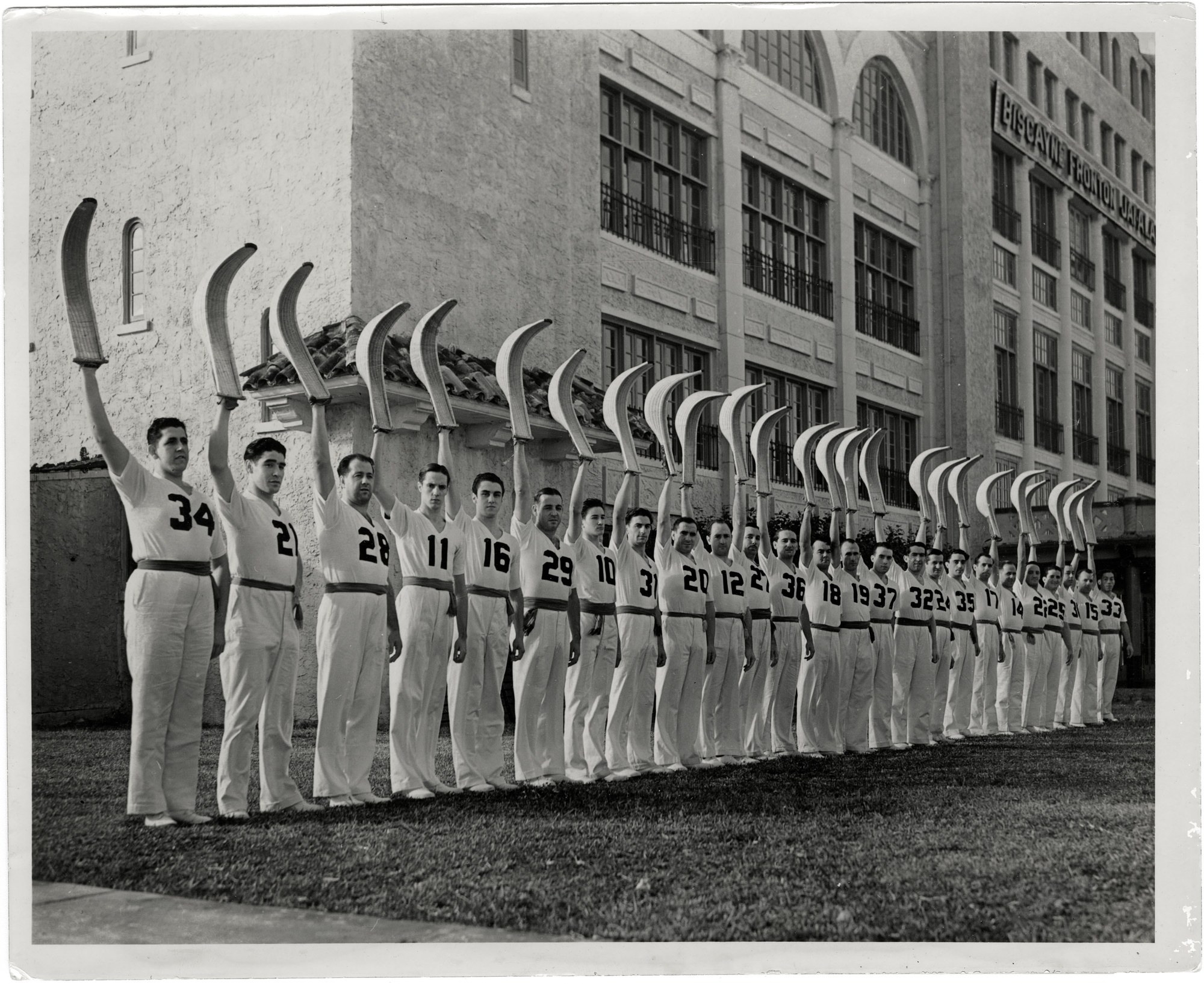 A long line of pelotaris in white uniforms, each holding a cesta raised overhead, standing outside the Biscayne Fronton Jai Alai building in Miami.