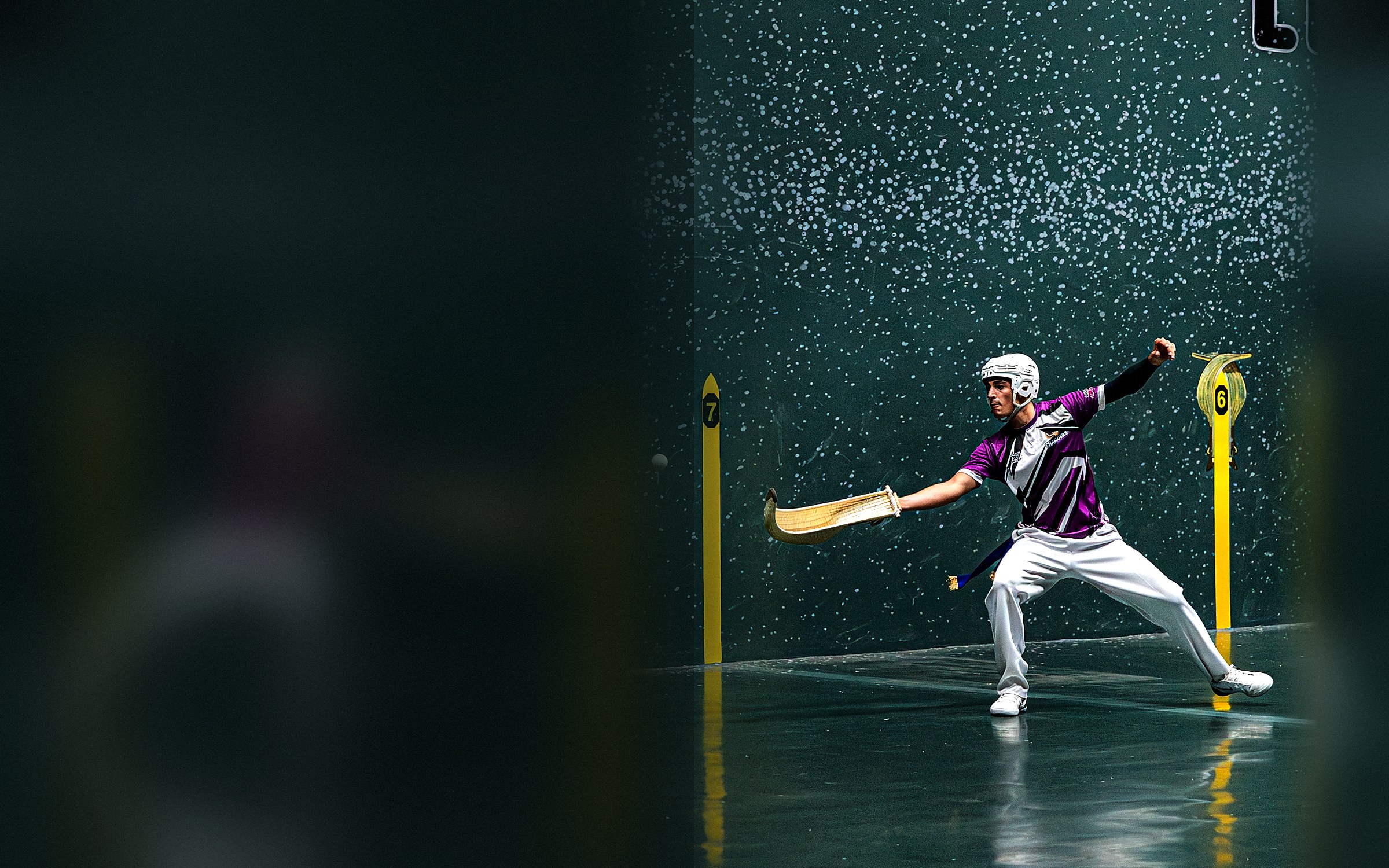 A World Jai Alai League pelotari with cesta extended, framed by yellow position markers against a ball-spattered fronton wall